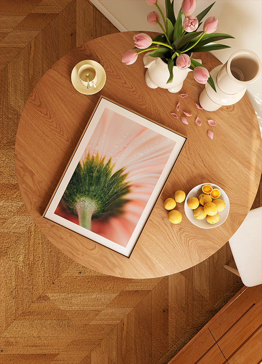 a wooden table with a framed picture of a flower, a bowl of fruit, and a vase of pink flowers.