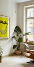 a cozy living room with a large window, a framed poster of a banana, a potted plant, and a comfortable armchair.