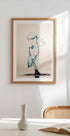 a framed artwork of a water droplet suspended in mid-air, with a blue and white color scheme. The artwork is displayed on a white wall above a table with a white vase and a book.