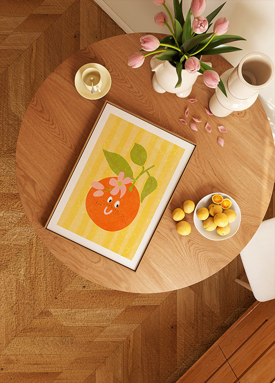 a wooden table with a framed picture of an orange, a bowl of lemons, a vase of pink tulips, and a candle on it.