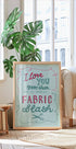 A framed poster with a handwritten message is displayed in a living room, with a potted plant and a bookshelf visible in the background.